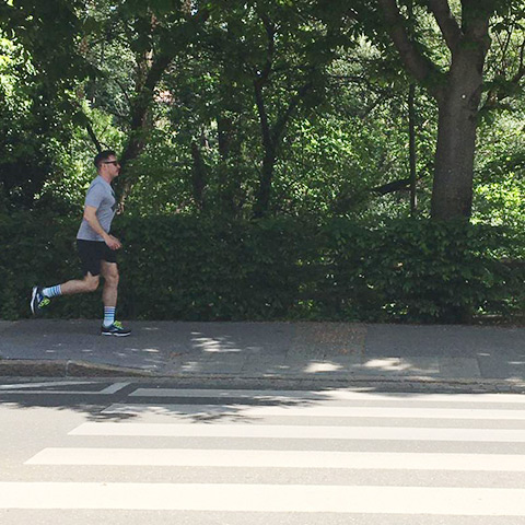 Eine Person joggt auf einem Gehweg neben einer Straße in einem üppigen, baumreichen Park. Sie trägt ein graues Hemd, schwarze Shorts und gestreifte Socken. Im Vordergrund ist ein Zebrastreifen zu sehen.