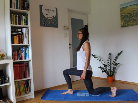 Eine Frau in einem weißen Tanktop und schwarzen Leggings macht einen Ausfallschritt auf einer blauen Yogamatte in einem Wohnzimmer mit Bücherregalen, einer Topfpflanze und einer Wanddekoration mit Fahrradmotiv.