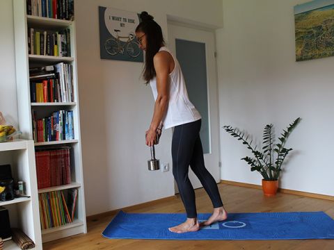 Eine Frau trainiert in einem Raum. Sie steht auf einer blauen Matte und hält in einer Hand ein Gewicht. Sie trägt ein weißes Tanktop und schwarze Leggings. Hinter ihr steht ein Bücherregal und an der Wand hängen ein Gemälde und ein Schild mit Fahrradmotiv. In der Nähe steht eine Topfpflanze.