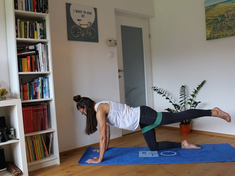 Eine Frau trainiert auf einer blauen Yogamatte mit einem Widerstandsband um ihre Beine. Sie befindet sich in einem Raum mit einem Bücherregal, Wandkunst und einer Topfpflanze. Ihr linkes Bein ist nach hinten ausgestreckt und ihr rechtes Knie liegt auf der Matte.