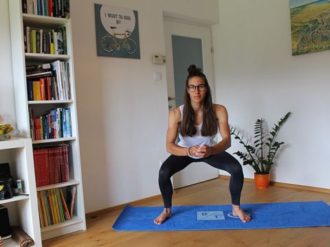 Eine Person mit langen Haaren und Brille macht Kniebeugen auf einer blauen Yogamatte in einem Wohnzimmer. Sie trägt ein weißes Tanktop und schwarze Leggings. Im Hintergrund sind Bücherregale, eine Pflanze und Wandkunst zu sehen.