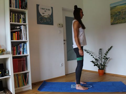 Eine Person steht in einem Raum auf einer blauen Yogamatte. Sie trägt ein weißes Tanktop und schwarze Leggings mit einem grünen Widerstandsband um die Oberschenkel. Im Hintergrund sind ein Bücherregal, ein Poster mit Fahrradmotiv und eine Pflanze zu sehen.