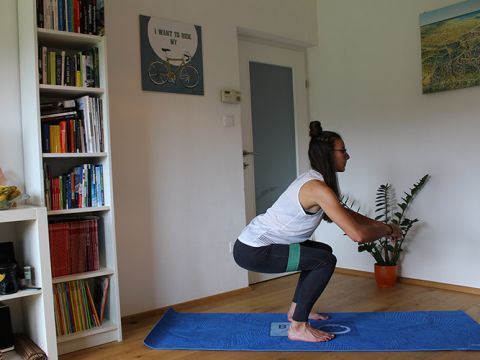 Eine Frau trainiert auf einer blauen Matte in einem Raum mit einem Bücherregal und einer Pflanze. Sie befindet sich in der Hocke, trägt ein weißes Tanktop, schwarze Leggings und über ihren Knien ein Widerstandsband. Zu sehen ist eine Wanddekoration mit Fahrradmotiv.