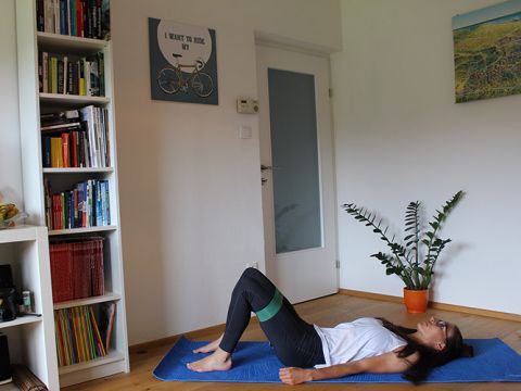 Eine Person liegt auf einer blauen Trainingsmatte in einem Wohnzimmer und führt eine Glute Bridge-Übung mit einem Widerstandsband um die Oberschenkel aus. Im Hintergrund sind ein Bücherregal und eine Topfpflanze zu sehen. An der Wand hängt ein Kunstwerk mit Fahrradmotiv.