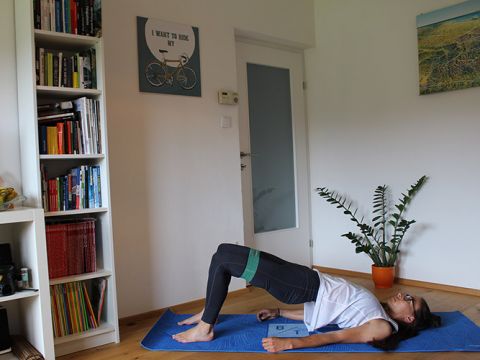 Eine Person macht eine Brückenpose auf einer blauen Yogamatte in einem Raum mit einem Bücherregal und einer Topfpflanze. Oben ist ein Bild mit einem Fahrrad und einem Text zu sehen. Die Person verwendet ein Widerstandsband an den Oberschenkeln und trägt eine Brille.