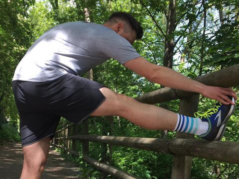 Eine Person streckt ihr Bein auf einem Holzzaun entlang eines belaubten, schattigen Pfades aus. Sie trägt ein graues Hemd, schwarze Shorts, gestreifte Socken und Turnschuhe und hat einen Fuß auf dem Zaun. Sonnenlicht fällt durch die grünen Bäume.