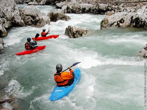 Drei Kajakfahrer befahren einen felsigen Wildwasserfluss. Sie tragen Helme und Schwimmwesten. Die Kajaks sind rot, blau und orange. Das Wasser ist schnell und die umliegenden Felsen sind schroff. Die Szene suggeriert Abenteuer und Teamwork.