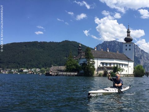 Eine Person fährt Kajak auf einem ruhigen See vor der Kulisse eines malerischen Gebäudes mit Kuppel und Türmen. Ein üppig grüner Berg steht unter einem klaren blauen Himmel und das Wasser spiegelt die lebendige Szene wider.