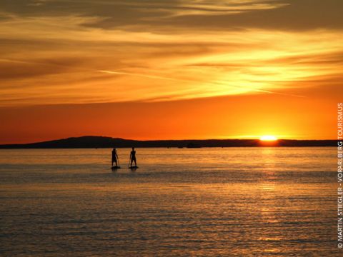 Zwei Personen paddeln auf einem ruhigen Gewässer während eines leuchtend orangefarbenen Sonnenuntergangs. In der Ferne zeichnet sich die Silhouette einer Insel ab, und dramatische Wolken betonen den Himmel.