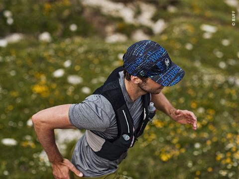 Eine Person läuft im Freien auf einer Wiese. Sie trägt eine blau gemusterte Mütze, ein graues T-Shirt und eine schwarze Trinkweste. Das Gelände ist mit gelben Blumen und Steinen übersät. Der Läufer ist konzentriert und bewegt sich zügig.