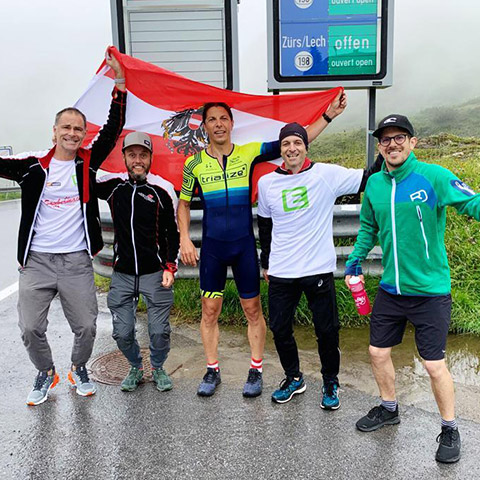 Fünf Menschen stehen lächelnd auf einer nassen Straße. Einer hält eine österreichische Flagge. Sie sind in Sportkleidung gekleidet und stehen vor einem nebligen Hintergrund. Im Hintergrund zeigt ein Schild die Richtung nach Zürs/Lech.
