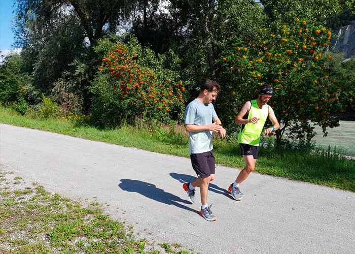 Zwei Menschen joggen auf einem sonnenbeschienenen Pfad in der Nähe eines Flusses. Sie tragen Sportkleidung; einer trägt ein hellblaues Hemd, der andere ein neongelbes Tanktop. Hinter ihnen stehen grüne Bäume mit orangefarbenen Früchten. Der Himmel ist klar und lässt einen hellen Tag erahnen.