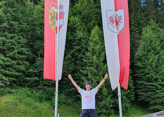Eine Person steht mit erhobenen Armen lächelnd zwischen zwei großen Flaggen mit Emblemen vor einer Kulisse aus dichten, grünen Bäumen. Sie trägt ein weißes Hemd, dunkle Shorts und eine Sonnenbrille.