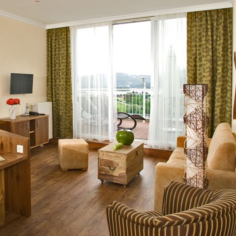 Hotelzimmer mit Blick auf eine Terrasse und einen See. Zur Ausstattung gehören ein beiges Sofa, zwei Würfelhocker, ein hölzerner Couchtisch mit einer grünen Vase, ein Fernseher an der Wand und eine dekorative Stehlampe. Vorhänge rahmen große Glastüren ein. Das Dekor ist warm und erdig.