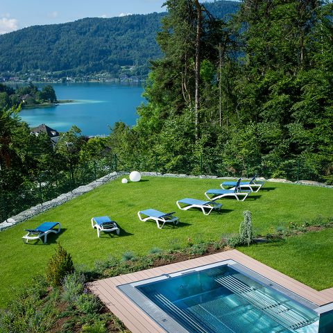 Eine malerische Aussicht bietet einen Rasen mit mehreren Liegestühlen neben einem modernen, rechteckigen Pool. Hinter dem Rasen liegt ein ruhiger blauer See, umgeben von üppigen grünen Hügeln und Bäumen unter einem klaren Himmel.