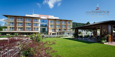 Vor einem strahlend blauen Himmel erhebt sich ein modernes Hotel mit zahlreichen Balkonen und großen Fenstern. Es ist von einer gepflegten Rasenfläche umgeben, auf der rechten Seite befindet sich ein kleiner Garten und ein schattiger Sitzbereich. Auf dem Gebäude prangt der Schriftzug „das balance“.