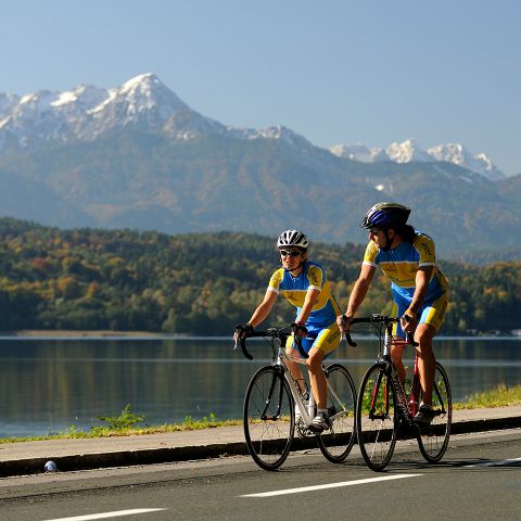 Zwei Radfahrer in passender blau-gelber Ausrüstung fahren mit ihren Rädern eine Uferstraße entlang. Den Hintergrund bilden schneebedeckte Berge unter einem klaren blauen Himmel. Über dem Wasser sind üppiges Grün und herbstliche Bäume zu sehen.