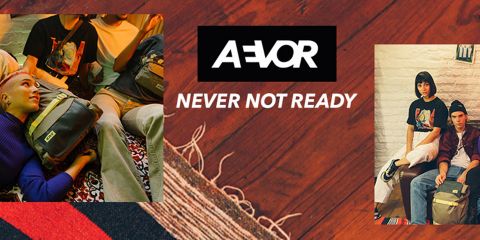 Drei Personen mit stylischen Rucksäcken und legerer Kleidung stehen in einem gemütlichen Raum. Auf einem gemusterten Hintergrund steht der Text „AEVOR NEVER NOT READY“.