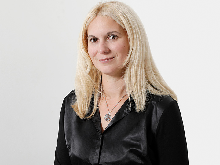 Eine Person mit langen blonden Haaren, die eine schwarze Bluse und eine Halskette mit rundem Anhänger trägt, steht vor einem schlichten weißen Hintergrund und lächelt leicht.