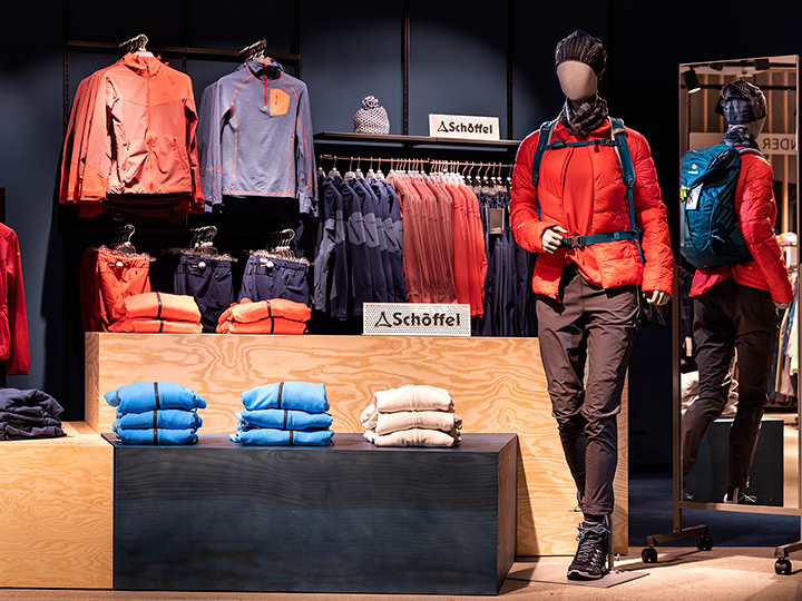 Schaufensterpuppe in Outdoor-Ausrüstung mit orangefarbener Jacke, schwarzer Hose, blauem Rucksack und Strickmütze. Auf den Ausstellungsregalen liegen gefaltete blaue und beige Kleidungsstücke und Kleiderbügel mit Jacken. Im Hintergrund ist das Schöffel-Logo zu sehen.