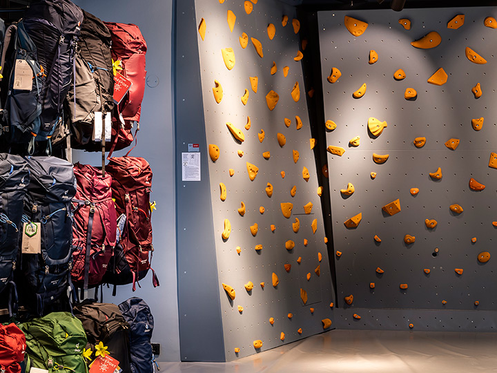 Indoor-Kletterwand mit verschiedenen orangefarbenen Griffen auf grauem Untergrund. Links daneben sind auf Gestellen mehrere Rucksäcke in unterschiedlichen Farben ausgestellt. Die Beleuchtung ist hell und der Boden poliert.