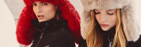 Zwei Menschen tragen große Pelzmützen, eine in leuchtendem Rot, die andere in Beige, und sind in dunkle Winterjacken gekleidet. Sie stehen dicht beieinander, während um sie herum sanft Schnee fällt und eine gemütliche, winterliche Szenerie schafft.