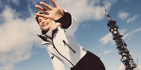 Eine Person in Winterkleidung streckt ihre Hand spielerisch in Richtung Kamera, während sie vor einem blauen Himmel mit Wolken steht. Auf der rechten Seite ist ein hoher Kommunikationsturm zu sehen.