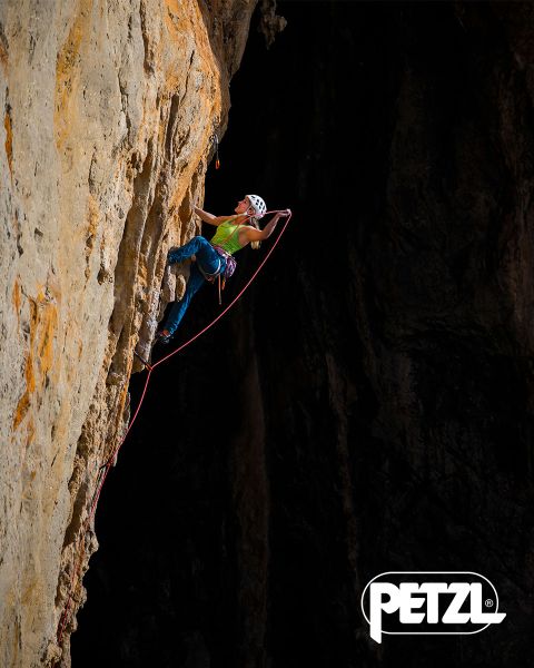Ein Kletterer in grüner Jacke und Helm erklimmt mit Eispickeln eine massive, hängende Eisformation. Schneebedeckte Felsen umgeben das Gebiet. Oben prangen die Worte „Experience the Difference“ und das Petzl-Logo.