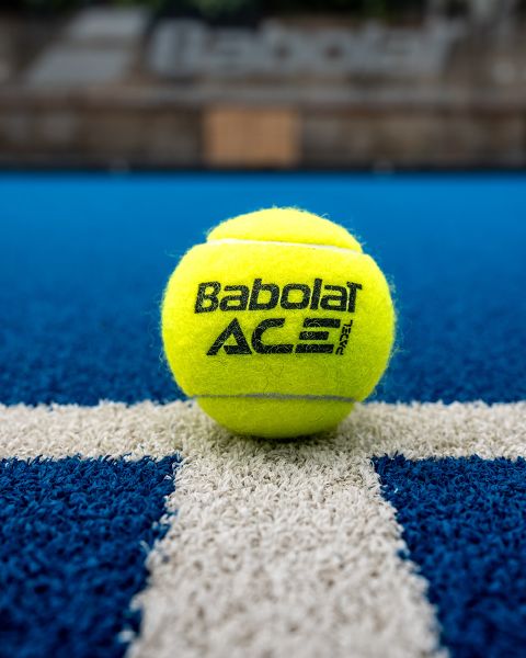 Ein gelber Babolat-Tennisball liegt auf einem blauen Tennisplatz, zentriert auf einer weißen Linie. Der Hintergrund ist unscharf und fokussiert das Markenlogo und die Textur des Balls.
