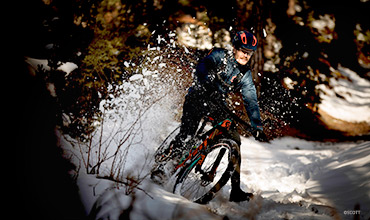 Eine Person in Winterkleidung fährt mit einem Mountainbike durch den Schnee in einem Wald und wirbelt dabei Schnee auf. Sonnenlicht fällt durch die Bäume und hebt die Radfahrszene und die winterliche Umgebung hervor.