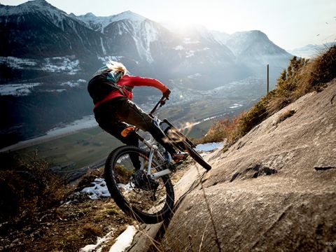 Ein Mountainbiker in einer roten Jacke fährt einen steilen, felsigen Abhang hinunter. Im Hintergrund sind schneebedeckte Berge und ein Tal unter einem teilweise bewölkten Himmel zu sehen. Helles Sonnenlicht beleuchtet den Radfahrer von hinten.