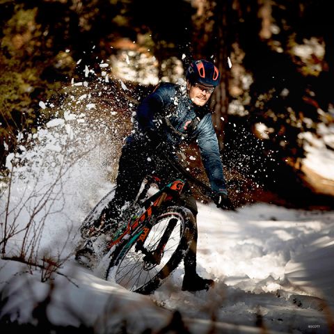 Eine Person auf einem Mountainbike rutscht durch verschneites Gelände und wirbelt dabei Schnee auf. Sie trägt einen Helm und Winterkleidung. Die Szene spielt in einem Waldgebiet mit verschwommenen Bäumen im Hintergrund.