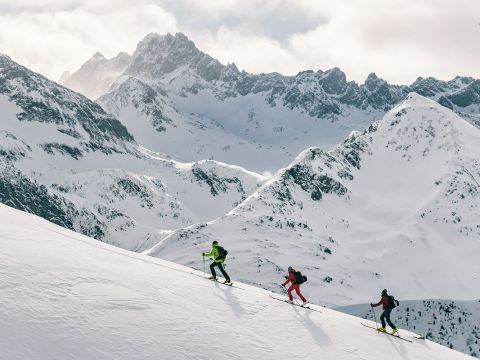 Drei Skifahrer in farbenfrohen Outfits fahren einen verschneiten Berghang hinauf, vor der Kulisse schroffer, schneebedeckter Gipfel. Der Himmel ist bedeckt, ein Hauch von Sonnenlicht bricht durch und wirft ein sanftes Licht auf die Szene.