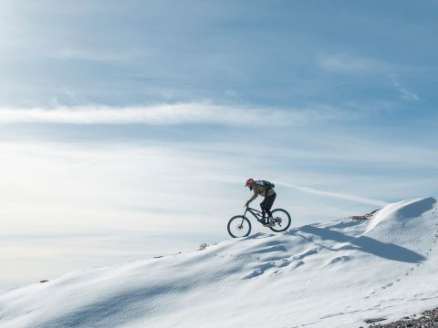 Eine Person mit Helm fährt unter einem klaren blauen Himmel mit dem Fahrrad über einen schneebedeckten Hügel. Im Schnee sind Reifenspuren sichtbar und die Landschaft ist weitläufig und ruhig, mit sanften Schatten, die von der Sonne geworfen werden.
