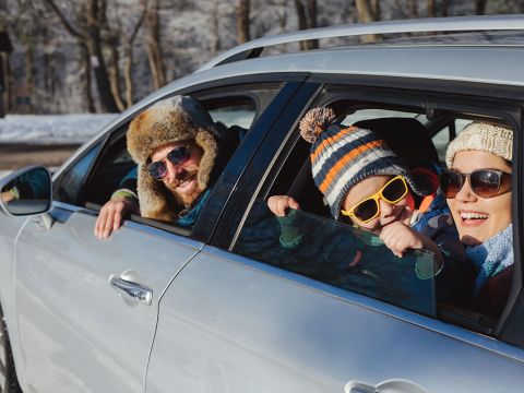 Eine Familie in Winterkleidung lehnt lächelnd aus einem silbernen Auto. Der Mann trägt eine Pelzmütze, das Kind eine Beanie und eine Sonnenbrille und die Frau eine Strickmütze und eine Sonnenbrille. Im Hintergrund sind schneebedeckte Bäume zu sehen.