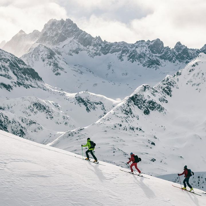 Drei Skifahrer fahren mit Skistöcken einen schneebedeckten Berghang hinauf, umgeben von einer atemberaubenden Aussicht auf schneebedeckte Gipfel und einen bewölkten Himmel. Die Szene vermittelt ein Gefühl von Abenteuer und der Schönheit der Winterlandschaft.