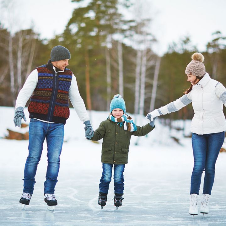 Eine dreiköpfige Familie läuft draußen auf einem zugefrorenen Teich Schlittschuh. Das Kind in der Mitte trägt einen grünen Mantel und eine blaue Mütze und hält an beiden Seiten die Hände eines Erwachsenen. Im Hintergrund sind hohe Bäume und schneebedeckter Boden zu sehen.