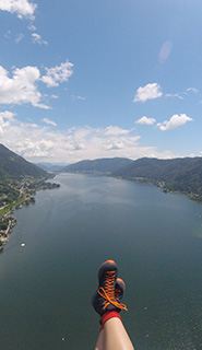 Luftaufnahme einer Person, die mit dem Gleitschirm über einen großen See fliegt. Ihre Füße sind zu sehen. Der See ist von Hügeln umgeben und am Himmel sind ein paar vereinzelte Wolken zu sehen. Die Szene ist ruhig und weitläufig.