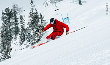 Ein Skifahrer in rotem Outfit fährt einen schneebedeckten Hang hinunter. Er ist nach vorne gebeugt und hält Skistöcke in der Hand. Im Hintergrund sind Bäume und ein Skilift zu sehen.