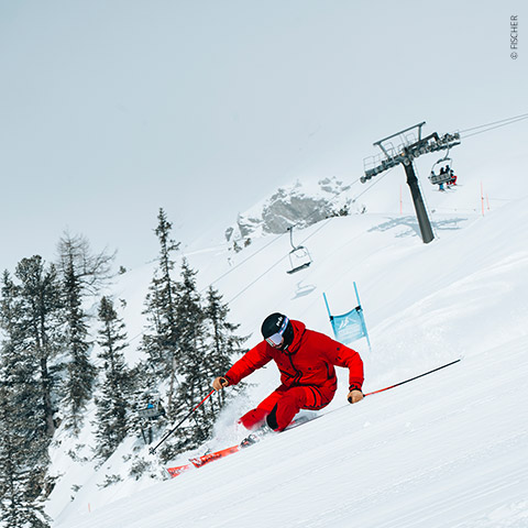 Ein Skifahrer in rotem Outfit fährt schnell einen schneebedeckten Hang hinunter und schneidet durch den Schnee. Im Hintergrund sind Sessellifte und Kiefern zu sehen, über ihm ein bewölkter Himmel.
