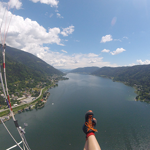 Eine Person fliegt mit dem Gleitschirm über einen malerischen See, der von Bergen und kleinen Städten am Seeufer umgeben ist. Der Blick von oben zeigt das blaue Wasser, üppige grüne Hügel und einen klaren Himmel. Die Füße des Gleitschirmfliegers sind in orangefarbenen Stiefeln zu sehen.