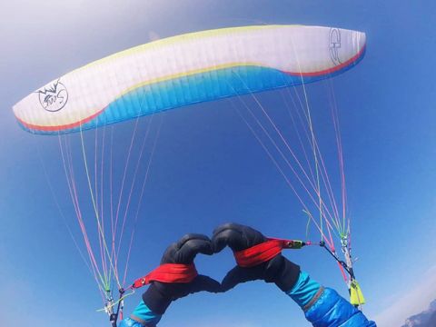Ein Gleitschirmflieger formt mit seinen behandschuhten Händen ein Herz, während er den bunten Gleitschirm über sich steuert. Der Himmel ist klar und strahlend blau, und das Gurtzeug des Gleitschirms weist leuchtende Linien auf.