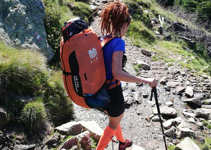 Eine Person mit einem großen orangefarbenen Rucksack und Wanderstöcken wandert einen felsigen Bergpfad entlang. Die Person trägt ein blaues Hemd, schwarze Shorts und orangefarbene Socken. Um sie herum sind grüne Grasflächen und verstreute Felsen.
