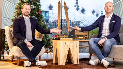 Zwei Männer sitzen in einem festlichen Raum mit einem geschmückten Weihnachtsbaum. Sie zeigen beide auf einen Holztisch, auf dem ein trophäenähnliches Objekt steht. Beide tragen legere Anzüge und lächeln. Im Hintergrund ist ein großes Fenster mit abstrakter Architektur zu sehen.