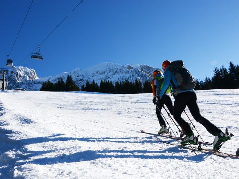 Zwei Skifahrer in Winterkleidung erklimmen mit Skiern und Stöcken einen schneebedeckten Hang. Im Hintergrund sind ein Skilift und ein klarer blauer Himmel zu sehen, und hinter immergrünen Bäumen erheben sich schneebedeckte Berge.