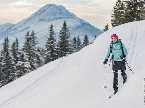 Eine Person, die in einer türkisfarbenen Jacke und einer rosafarbenen Mütze Ski auf frischem Schnee fährt. Im Hintergrund sind schneebedeckte Bäume und ein majestätischer Berggipfel zu sehen.