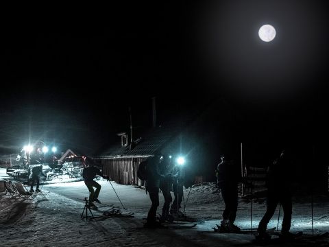 Skifahrer mit Stirnlampen fahren bei Vollmond einen schneebedeckten Hang hinauf, in der Nähe einer Hütte mit Beleuchtung. Die Szene ist dunkel, erhellt durch Mondlicht und künstliches Licht, was eine heitere und abenteuerliche Atmosphäre schafft.