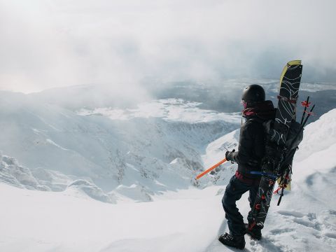 Ein Snowboarder mit Helm und Schneeausrüstung steht auf einem schneebedeckten Berghang, hält einen Eispickel und blickt auf eine weite, neblige Landschaft. Auf seinem Rücken ist ein Snowboard festgeschnallt. Die Szene ist hell und winterlich.