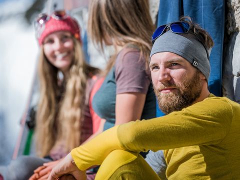Drei Personen in Outdoor-Kleidung sitzen und unterhalten sich. Im Fokus steht ein bärtiger Mann in Gelb, der eine Sonnenbrille auf dem Kopf trägt. Eine Frau mit einer roten Beanie und eine andere in einem grauen Hemd lächeln im Hintergrund, leicht unscharf.