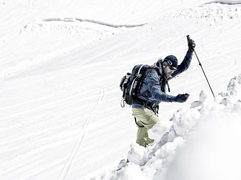 Eine Person in Winterkleidung erklimmt einen schneebedeckten Berghang und stützt sich dabei auf einen Stock. Im Hintergrund ist eine weite Schneefläche zu sehen, der Himmel ist nicht sichtbar. Die Szene vermittelt ein Gefühl von Abenteuer und Entschlossenheit.